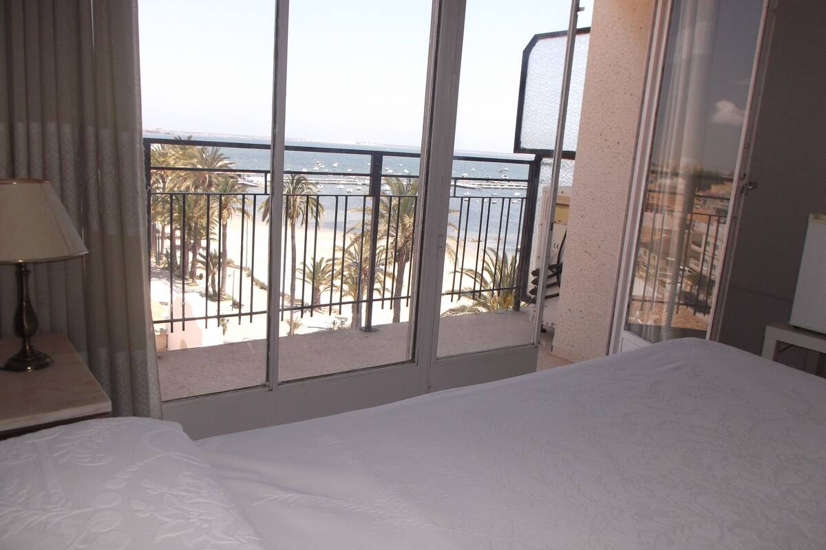A hotel room with a bed, a nightstand with a lamp, and a balcony with a view of the beach and ocean.