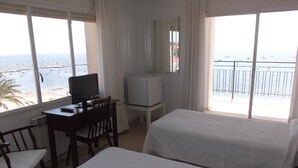 Ein Hotelzimmer mit zwei Betten, einem Schreibtisch mit Computer, einem Sessel und einem Fernseher. Das Zimmer verfügt über große Fenster mit Blick auf das Meer und Boote.