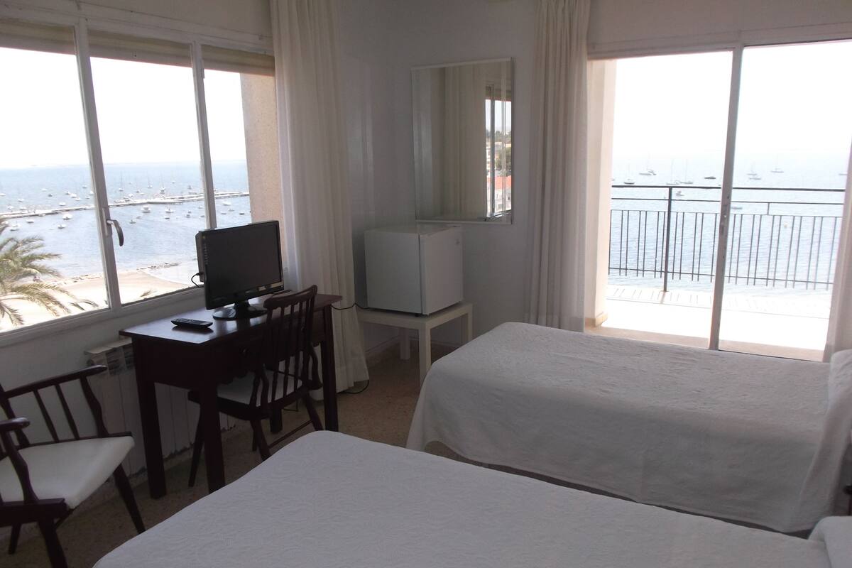 A hotel room with two beds, a desk with a computer, a chair, and a TV. The room has large windows with a view of the sea and boats.