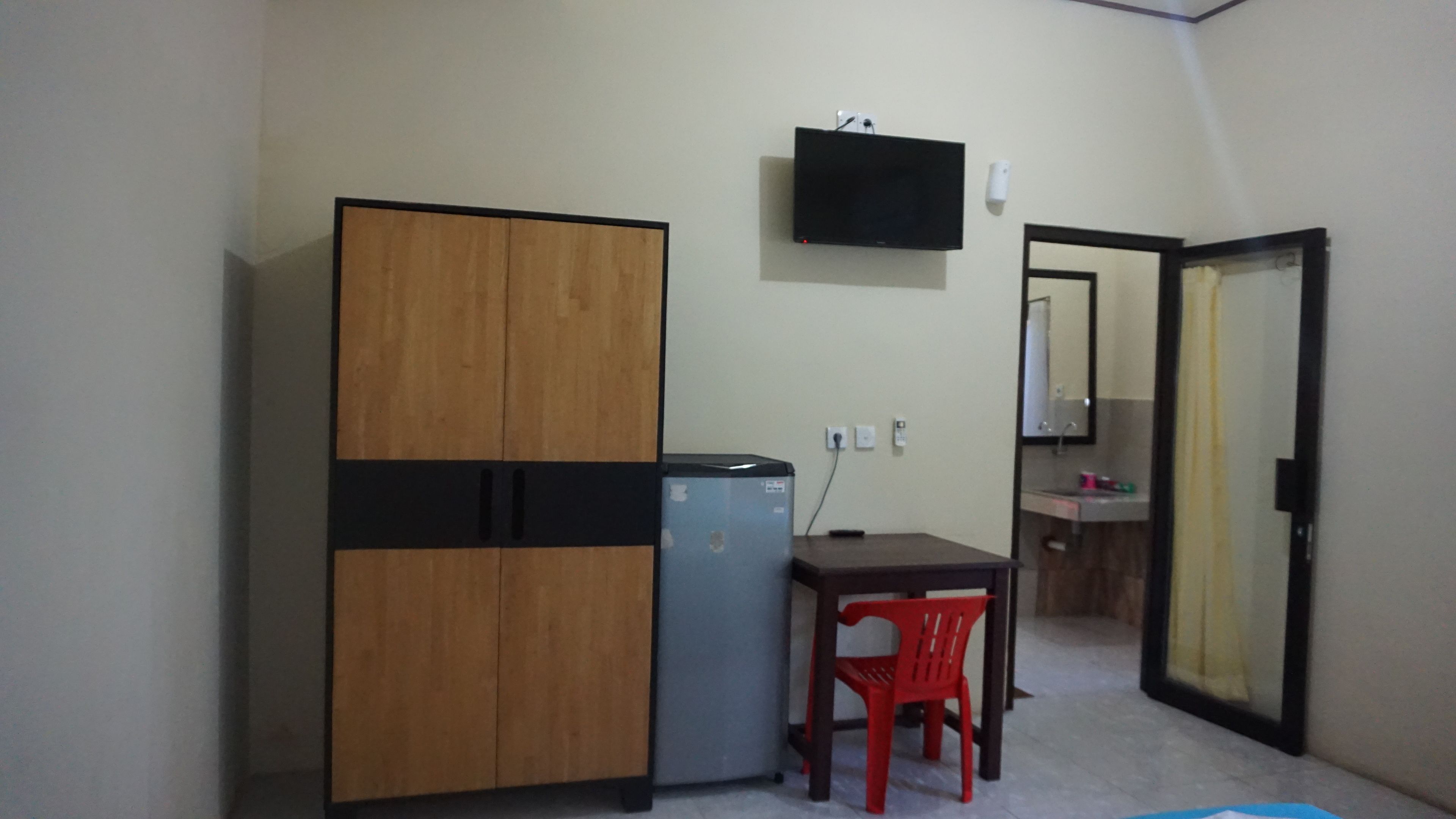 Una habitación pequeña con un armario de madera, un refrigerador pequeño, un escritorio con una silla roja, un televisor montado en la pared y un baño visible a través de una puerta abierta.
