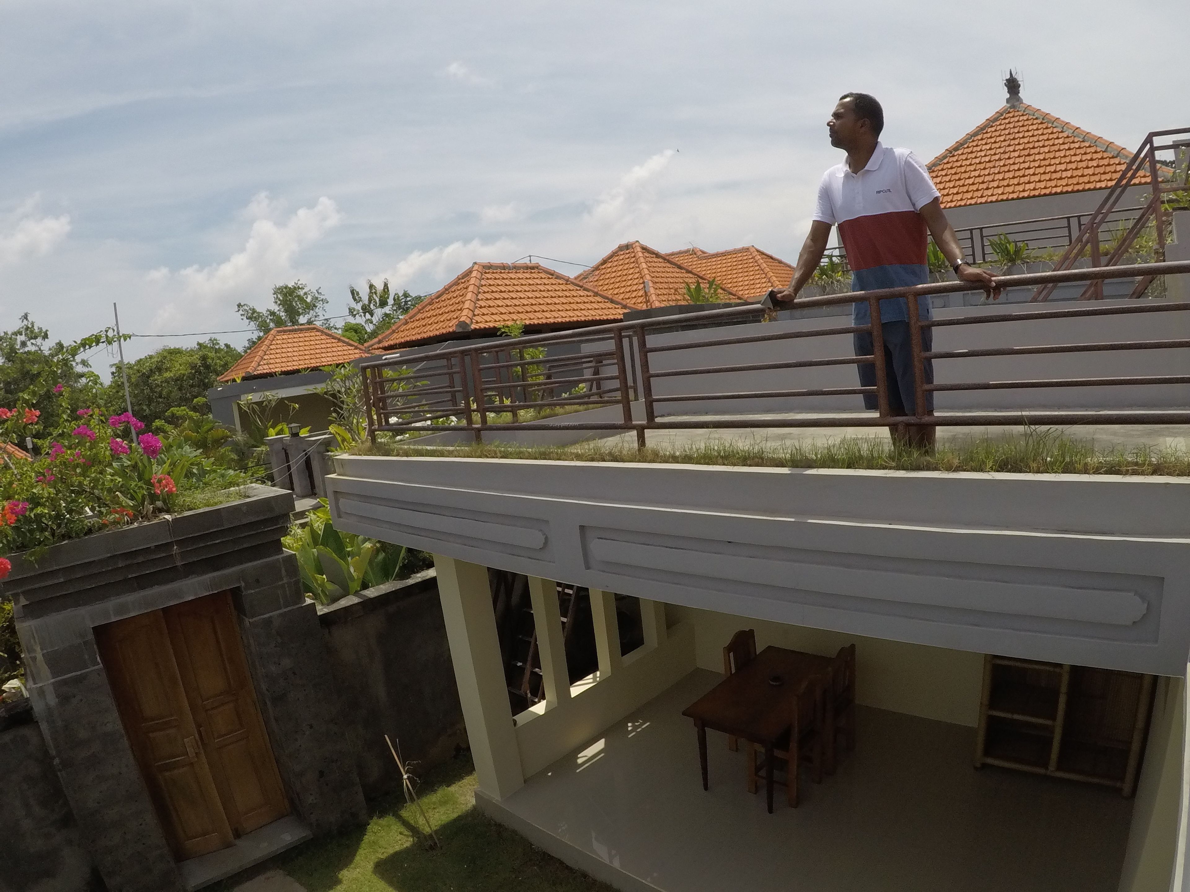 Un hombre está de pie en un balcón con vistas a casas de tejados de terracota y un jardín lleno de flores rosadas.