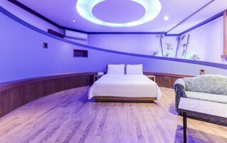 A modern bedroom with a circular ceiling light, a bed with white linens, a wooden floor, and a green armchair.