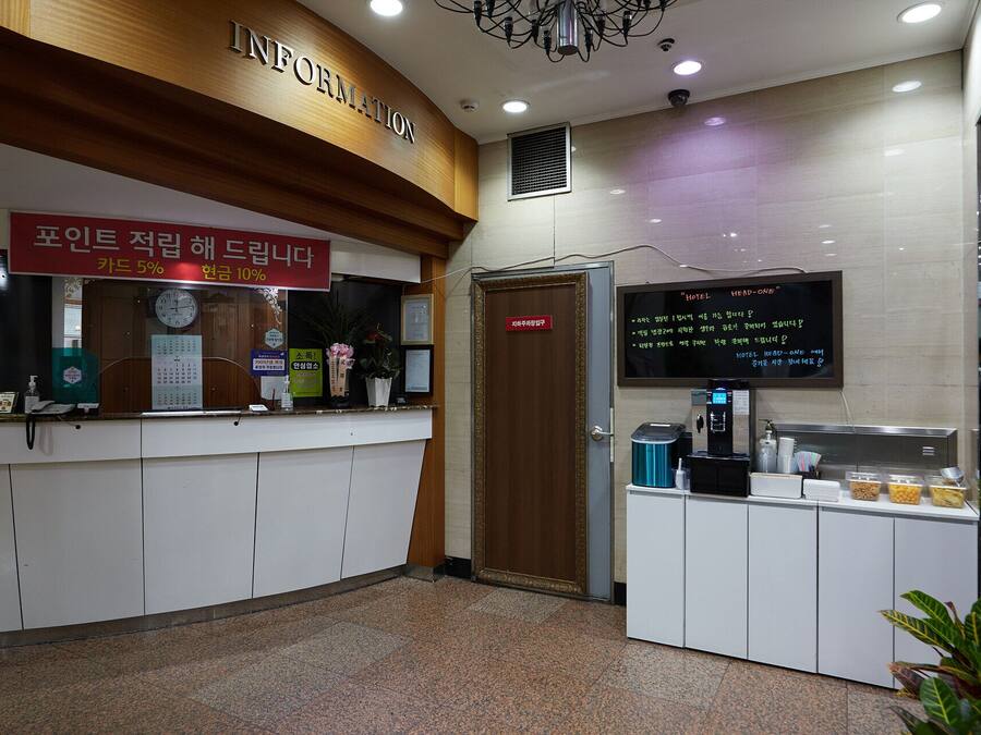 A hotel reception area with a wooden reception desk, a digital information board, a coffee machine, and a small plant.