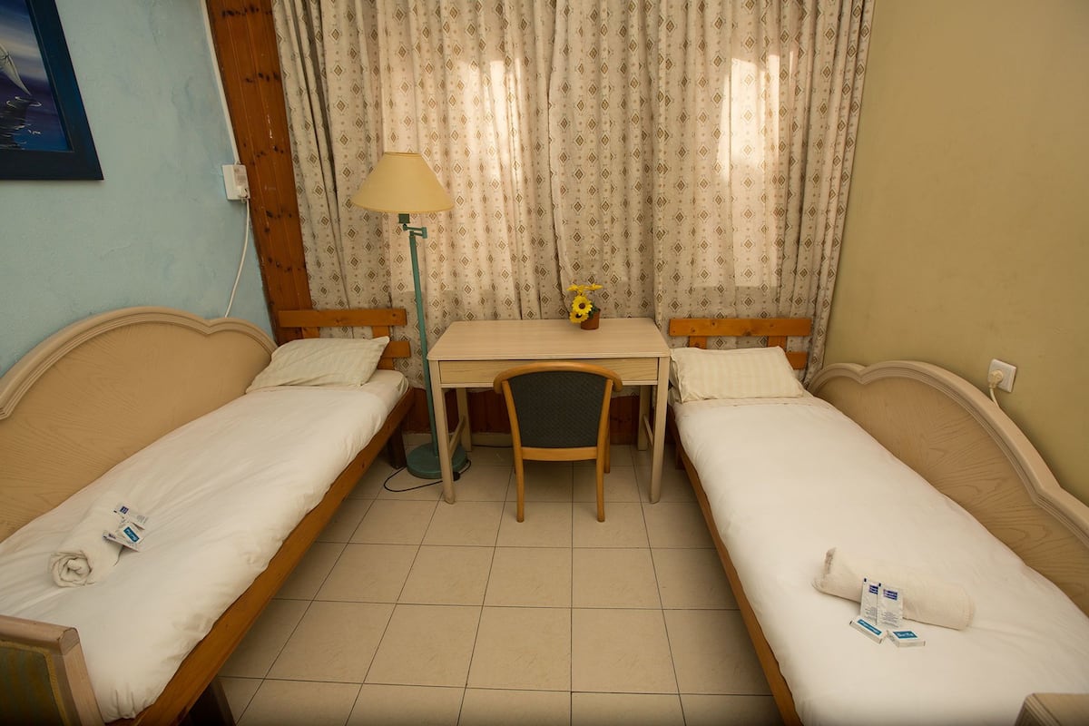 A room with two single beds, a small desk with a chair, a lamp, and a window with patterned curtains.