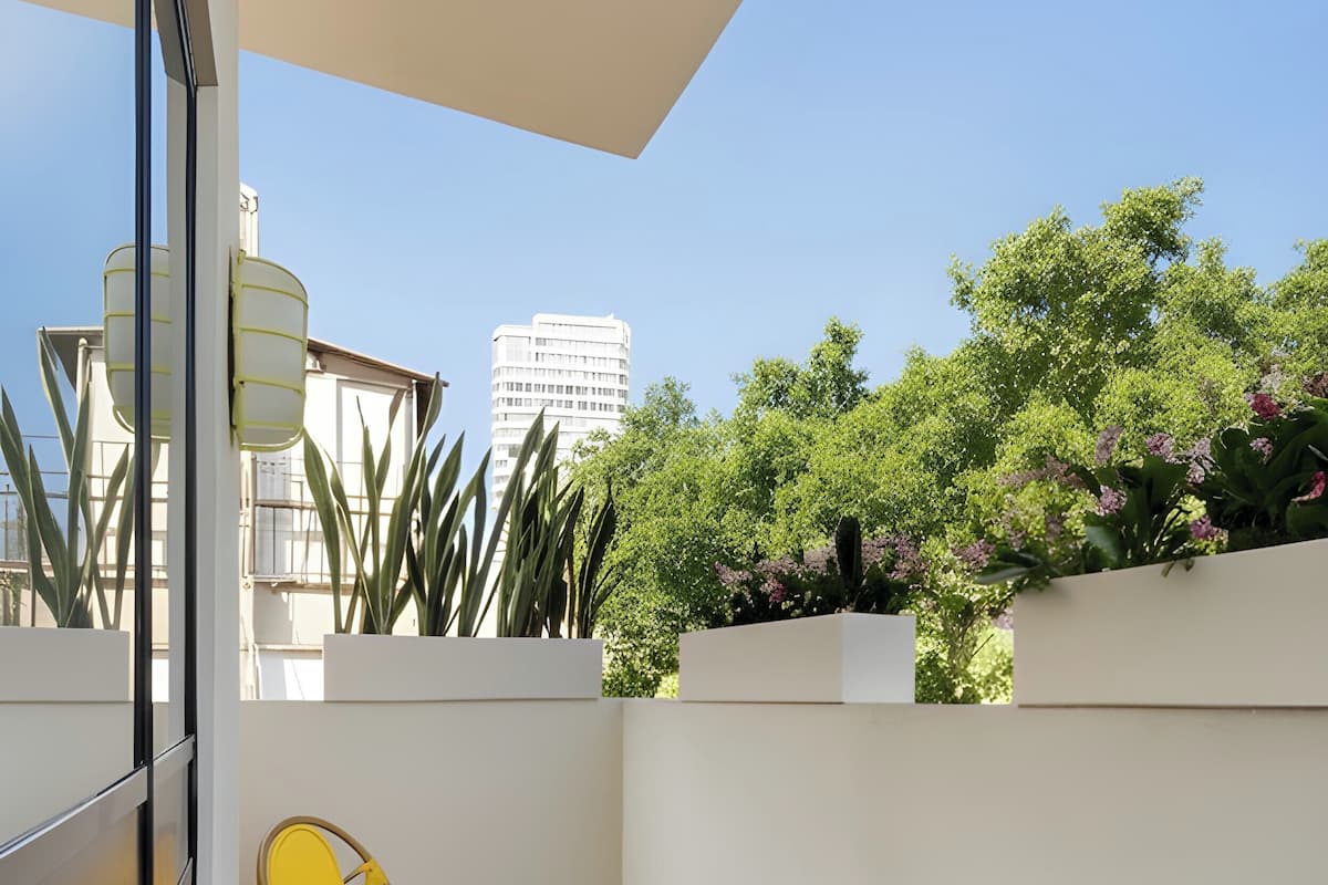 Un balcón con una silla de mimbre y una mesita pequeña, con vistas a un jardín con árboles y un edificio al fondo.