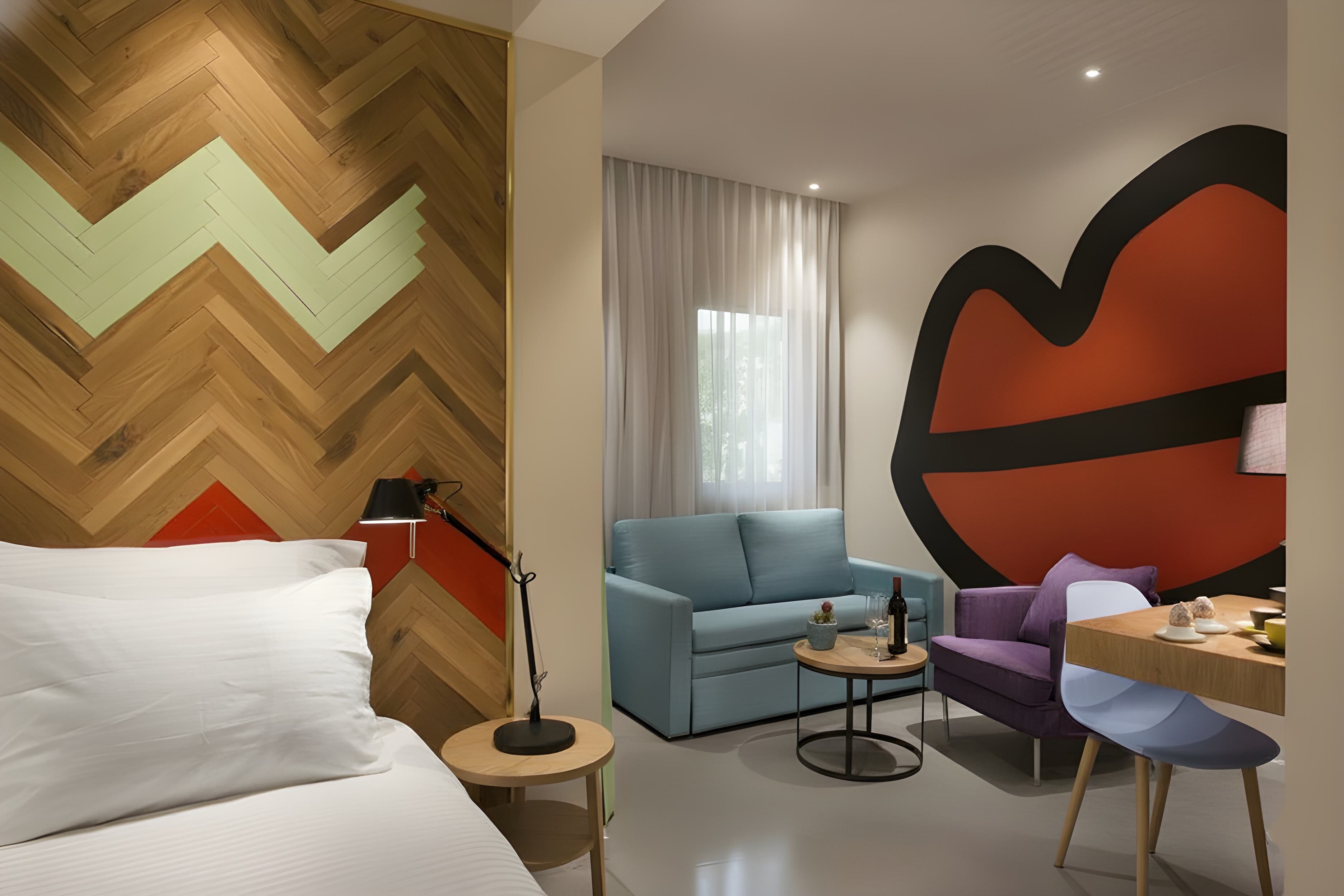 Een moderne hotelkamer met een houten visgraatvloer, een bed met witte lakens, een blauwe bank, een paarse fauteuil, een klein rond tafeltje met een lamp, en een groot abstract kunstwerk aan de muur.