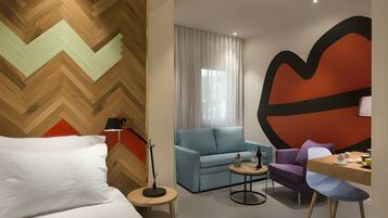 Een moderne hotelkamer met een houten visgraatvloer, een bed met witte lakens, een blauwe bank, een paarse fauteuil, een klein rond tafeltje met een lamp, en een groot abstract kunstwerk aan de muur.