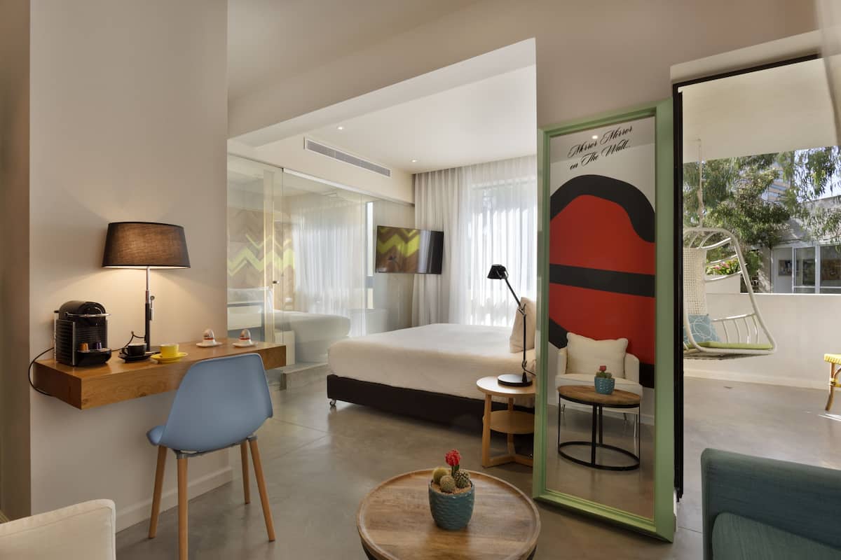 Una habitación de hotel moderna con una cama, un escritorio con lámpara, una silla, una mesita con un cactus y un espejo grande.