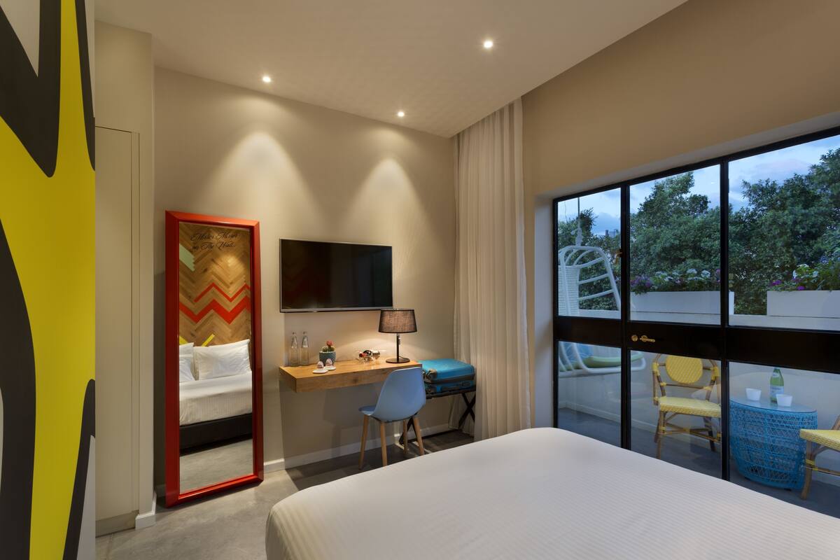 Habitación de hotel con cama, escritorio, silla, televisión, ventana con vista a árboles y espejo.