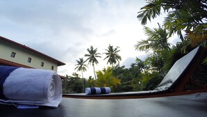 Zona junto a la piscina con sillones de descanso y toallas, palmeras y un edificio al fondo.