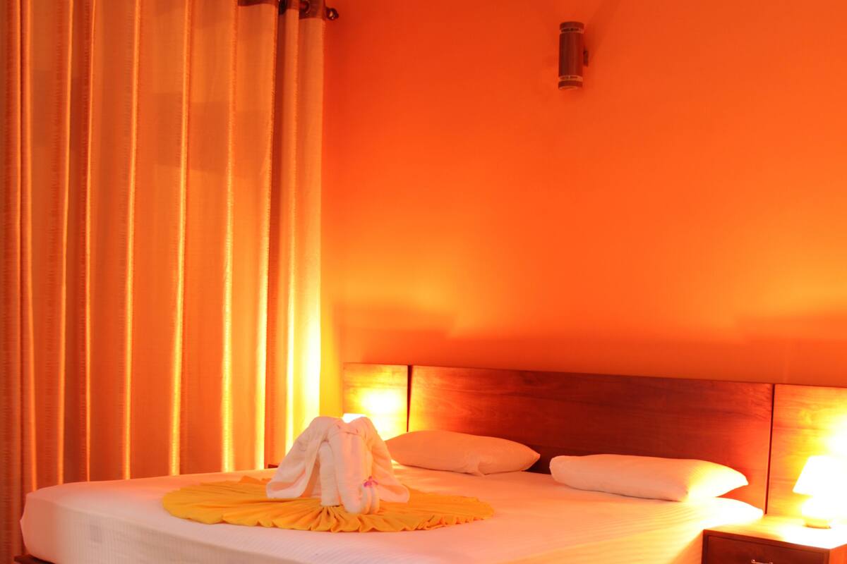 Una habitación de hotel con una cama de madera, ropa de cama blanca, una alfombra roja y una mesita de noche con una lámpara.
