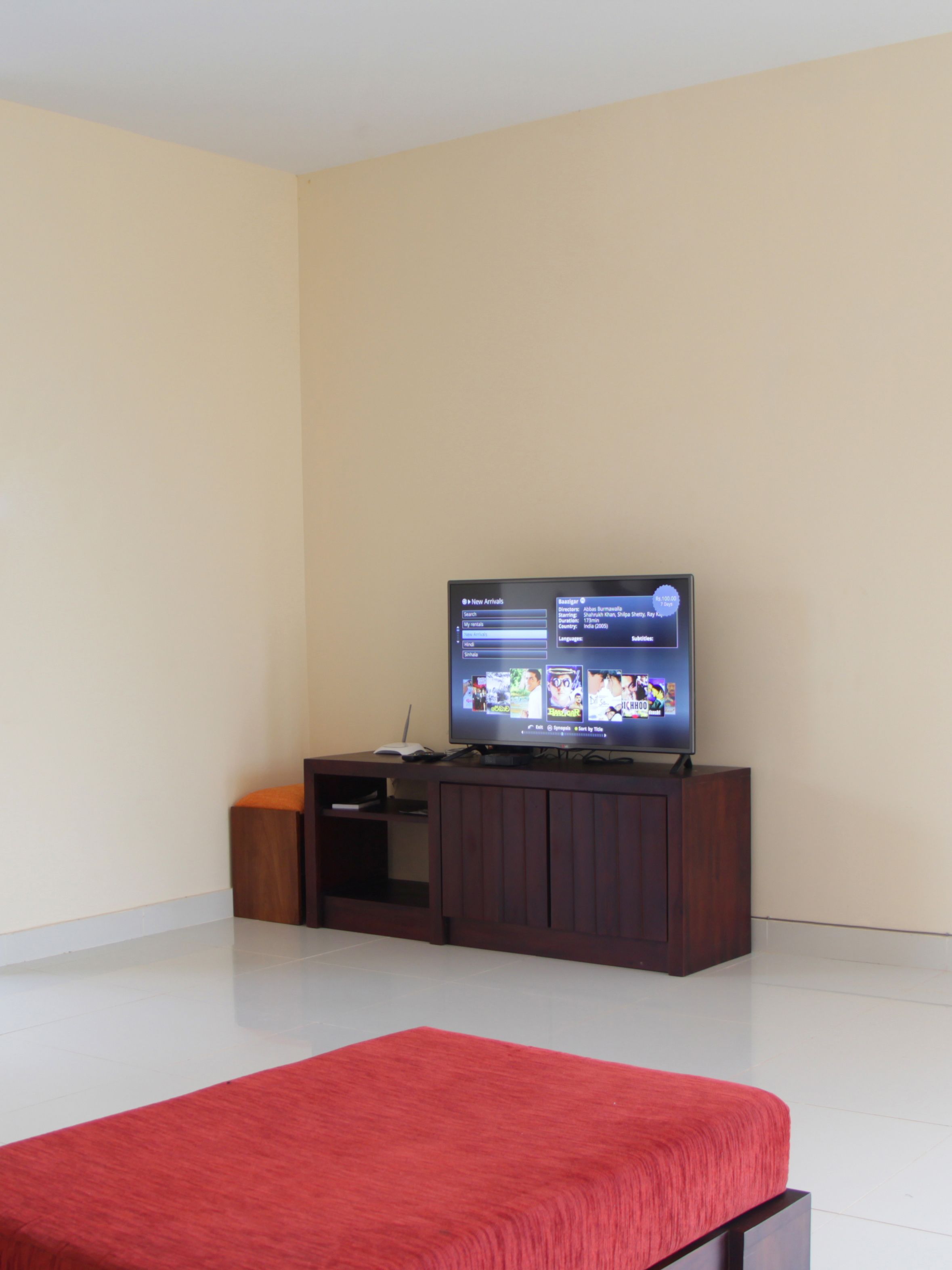 A room with a flat-screen TV on a wooden stand, a red bedspread, and a white tiled floor.