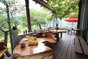 Una terraza de madera al aire libre con mesas y bancos, rodeada de árboles y con vistas a un paisaje montañoso.