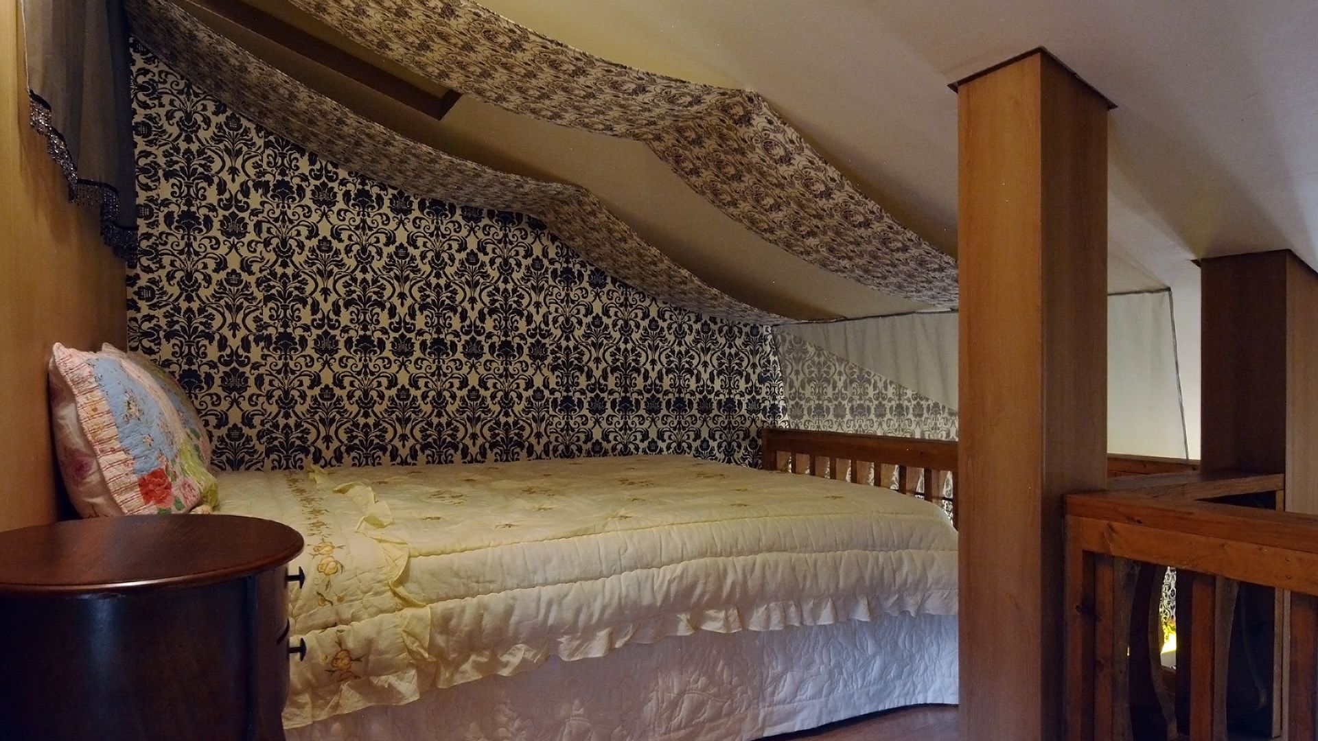 Un dormitorio con papel tapiz estampado, una cama con cubrecama floral, cabecera de madera y mesita de noche con lámpara.