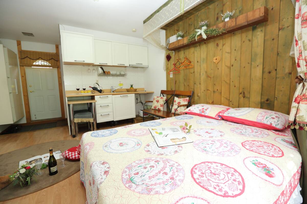 A bedroom with a wooden headboard, a bed with a floral quilt, a bedside table with a book, and a small table with a bottle and glasses.