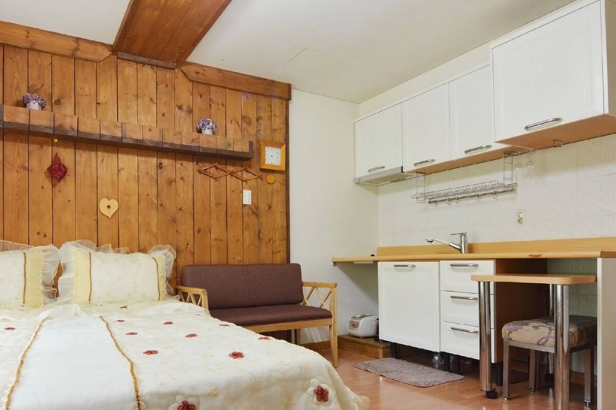 A bedroom with a bed, a wooden headboard, a brown sofa, and a kitchenette with white cabinets and a sink.