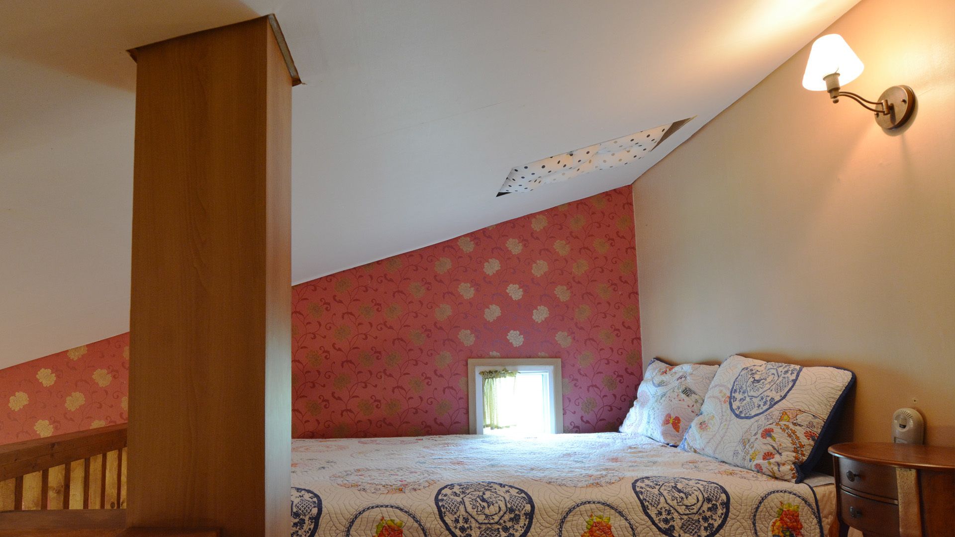 Een slaapkamer met een schuine plafond, een houten trap, een bed met beddengoed met een patroon, een nachtkastje en een wandlamp.