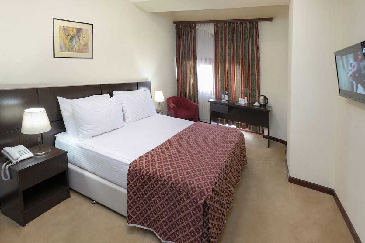 Habitación de hotel con una cama, una mesita de noche con teléfono, un escritorio con silla, un televisor y una ventana con cortinas.