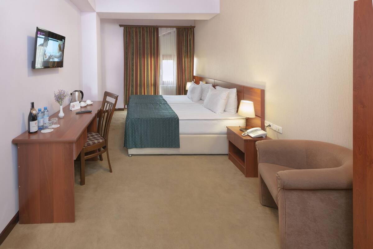 Una habitación de hotel con cama, mesita de noche, silla, escritorio, televisor y cortinas.