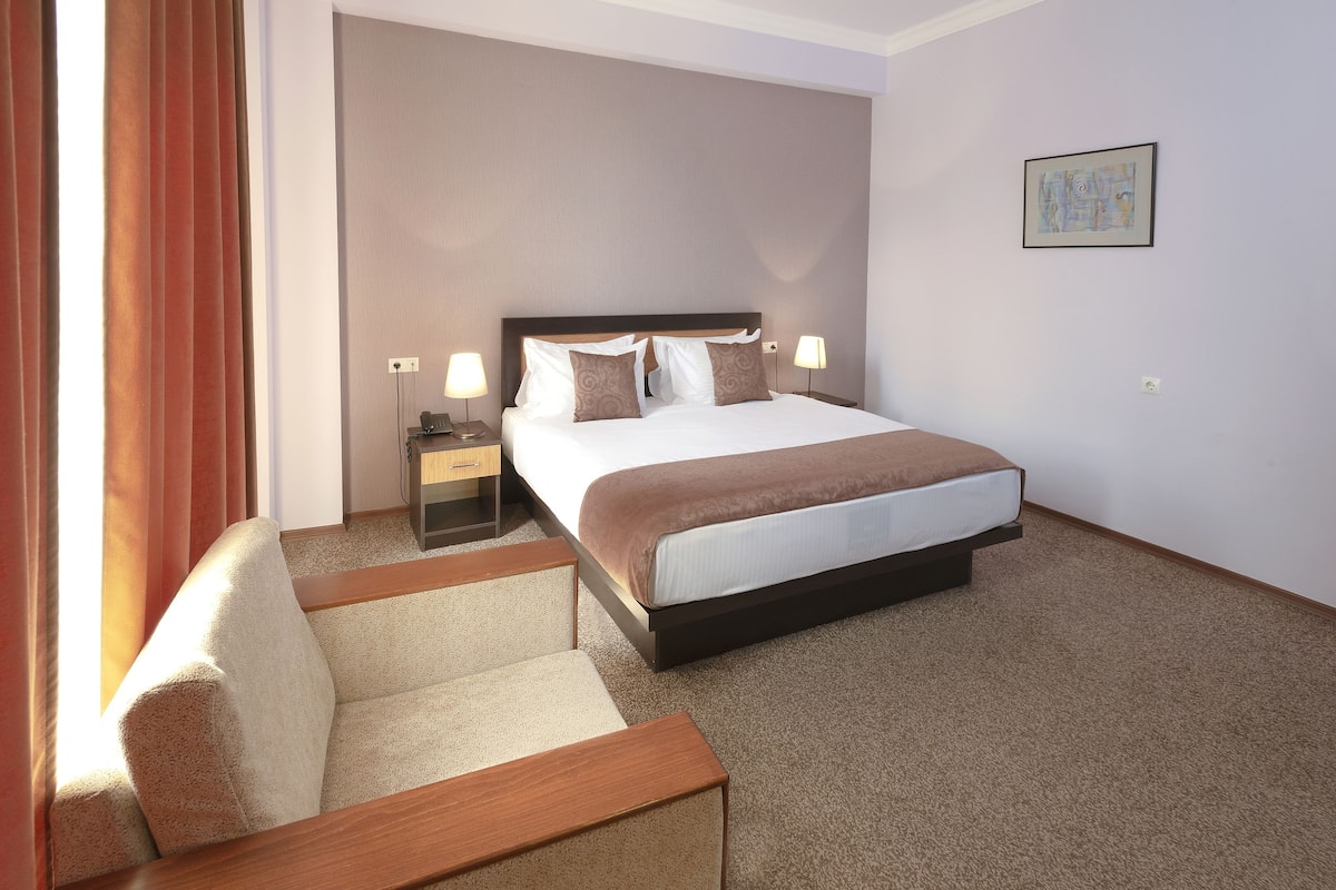 Una habitación de hotel con una cama grande, mesitas de noche, una silla y un cuadro en la pared.