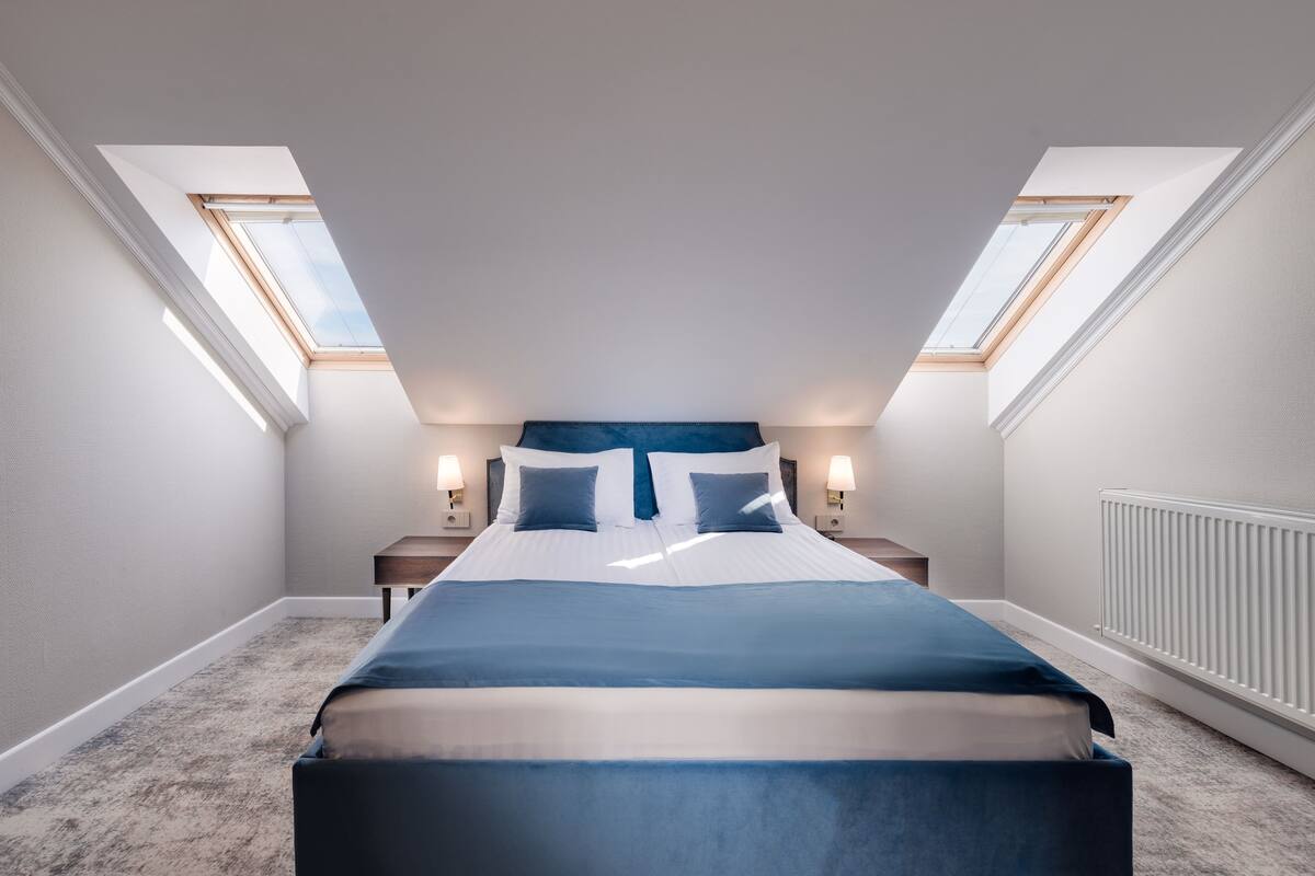 Un dormitorio con tragaluz, cama con ropa de cama azul y mesita de noche con lámpara.