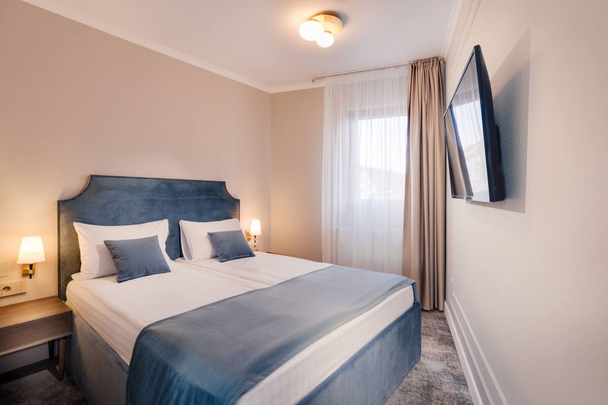 Habitación de hotel con una cama grande, una mesita de noche con lámpara, un televisor de pantalla plana en la pared y una ventana con cortinas.