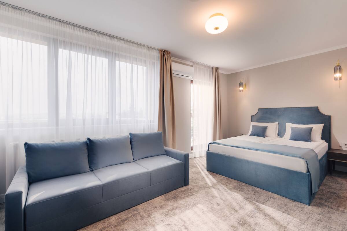 Habitación de hotel con una cama grande, un sofá y una ventana con cortinas traslúcidas.