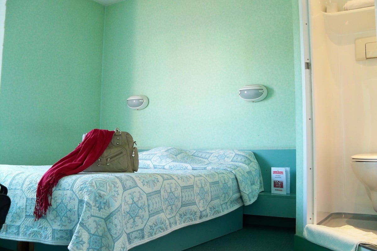 Een kleine kamer met een enkel bed, een groene muur, een wit toilet en een rode tas op het bed.