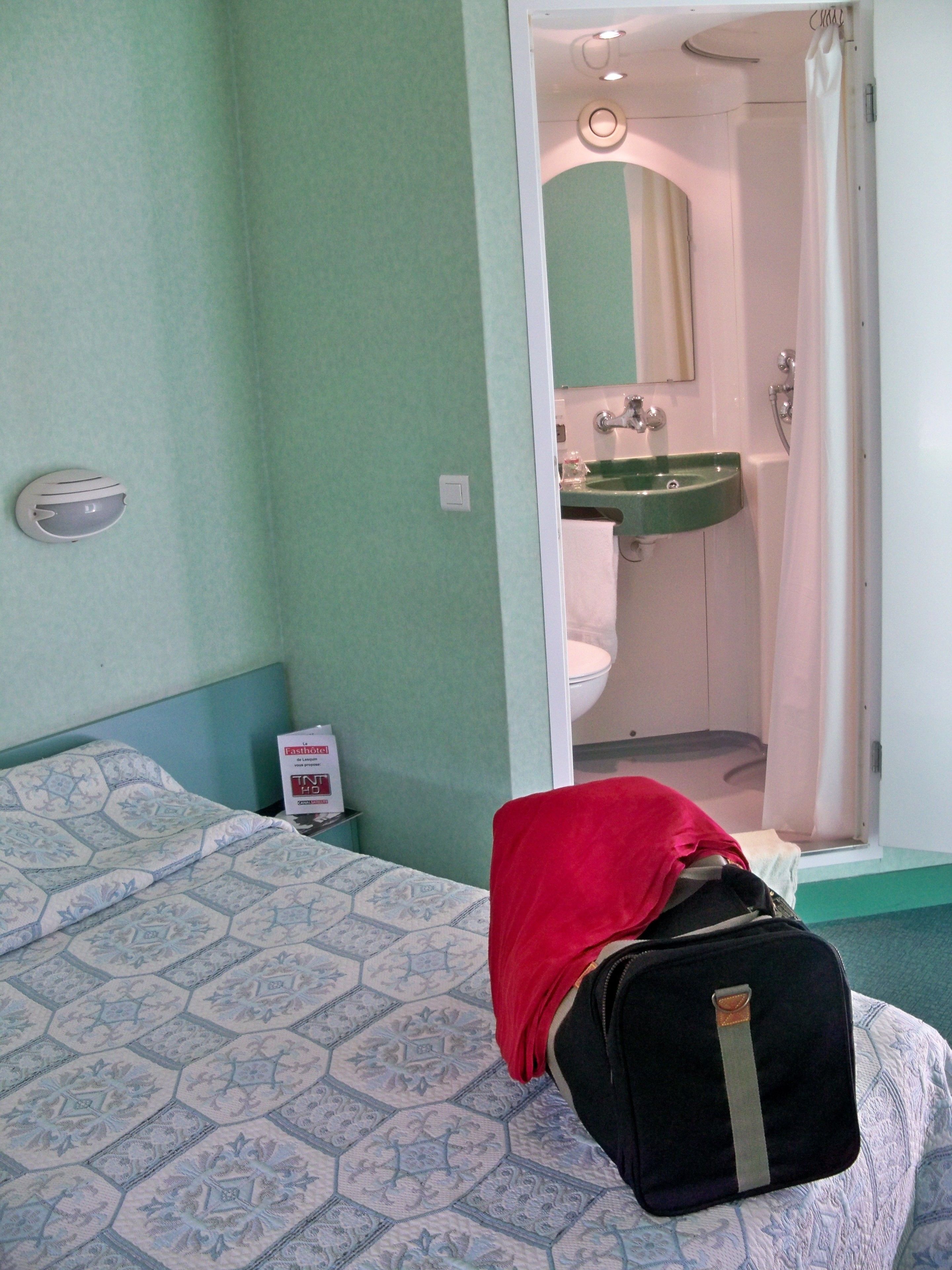 Habitación de hotel con cama, baño a la vista a través de una puerta abierta y mesita de noche con lámpara.