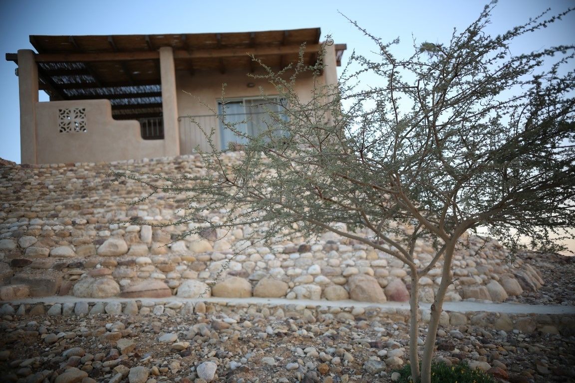Un bâtiment à la façade en pierre et au toit plat, entouré d’un terrain rocheux et d’une végétation clairsemée.