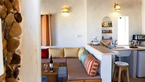 A cozy kitchen with a stone wall, wooden beams, a built-in bench with cushions, a wooden stool, and a countertop with a sink and various kitchen items.