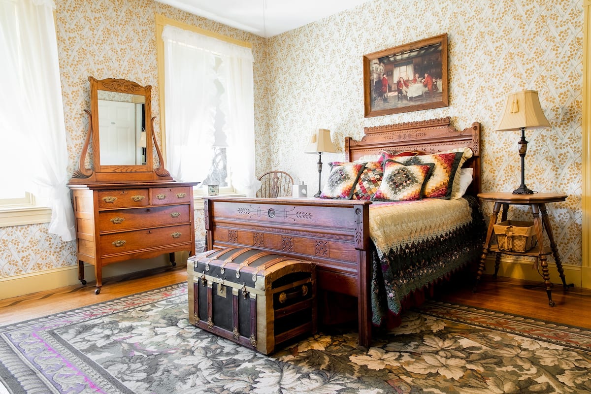 A bedroom with a wooden bed, a dresser, a trunk, a mirror, and a painting on the wall.