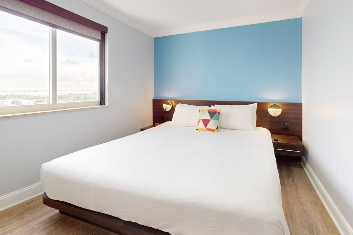 A hotel room with a large bed, a window with blinds, a blue accent wall, and a wooden headboard.