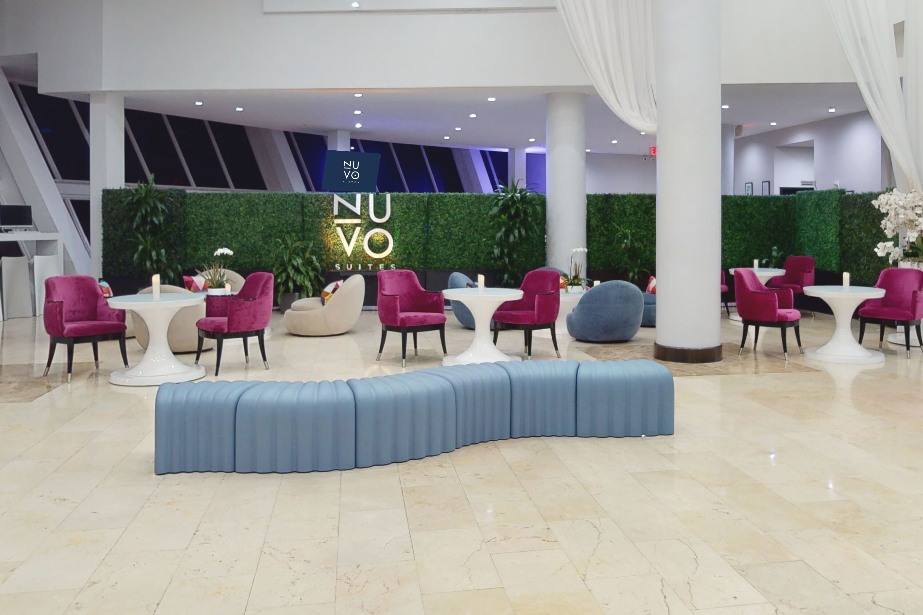 A modern hotel lobby with a reception area featuring a green wall, a sign reading "NUVO SUITES," and a seating arrangement with round tables and chairs.