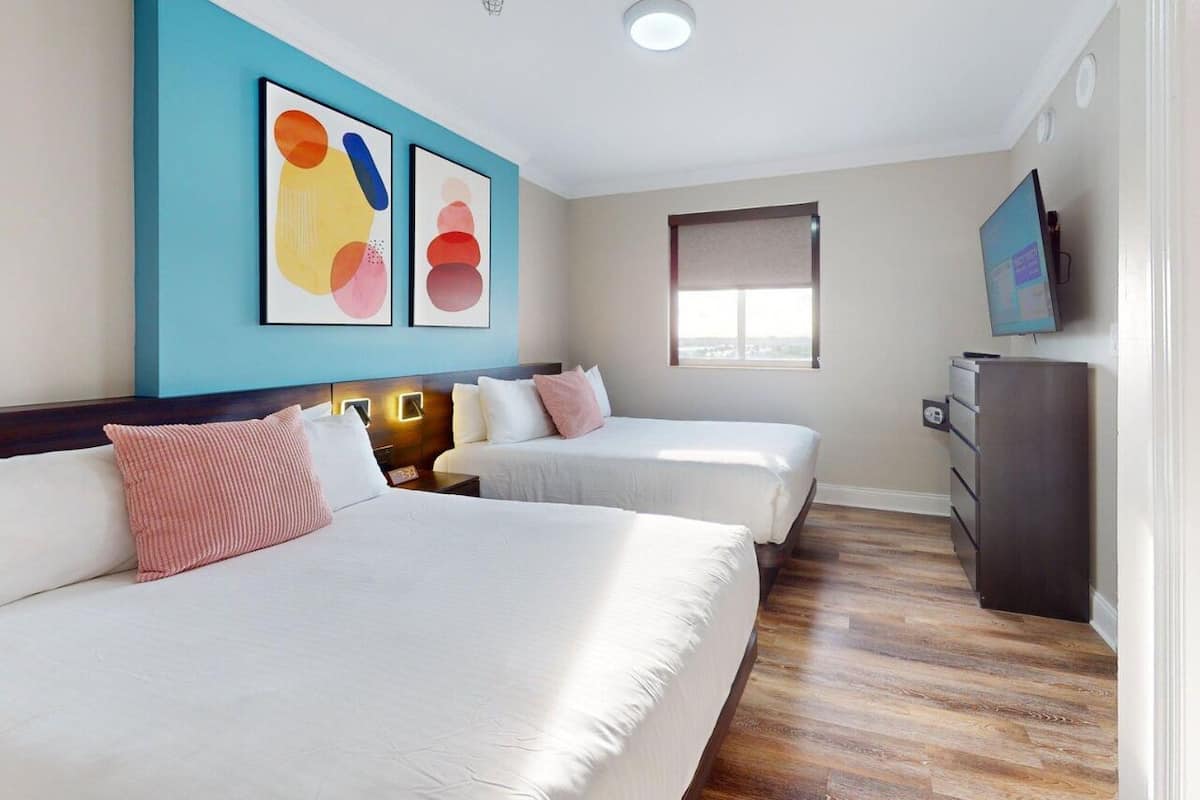 A hotel room with two beds, a desk, a TV, and artwork on the wall.