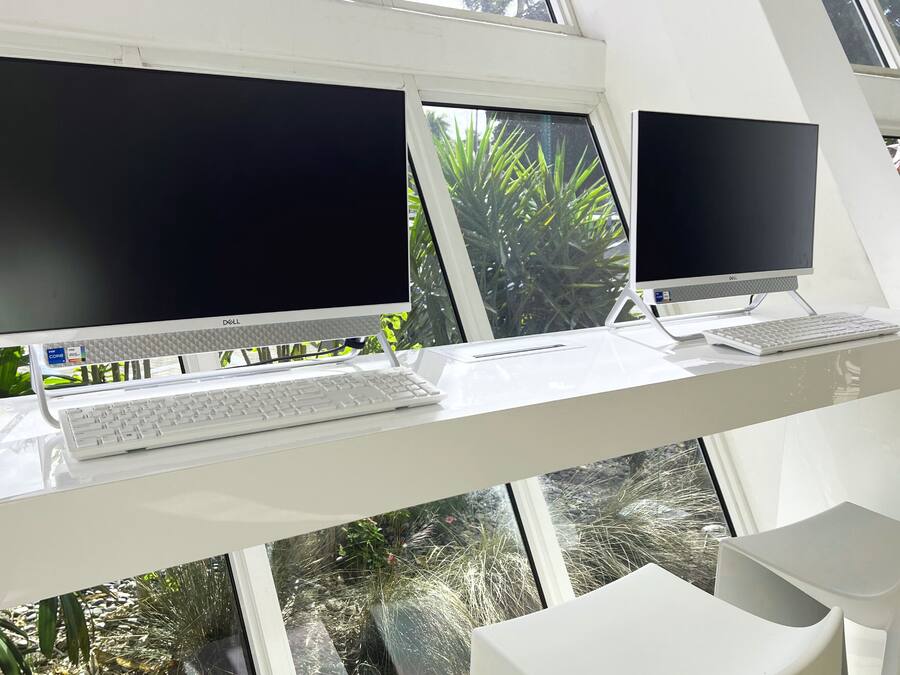 Una oficina moderna con dos monitores Dell, un escritorio blanco y vistas a zonas verdes a través de amplios ventanales.
