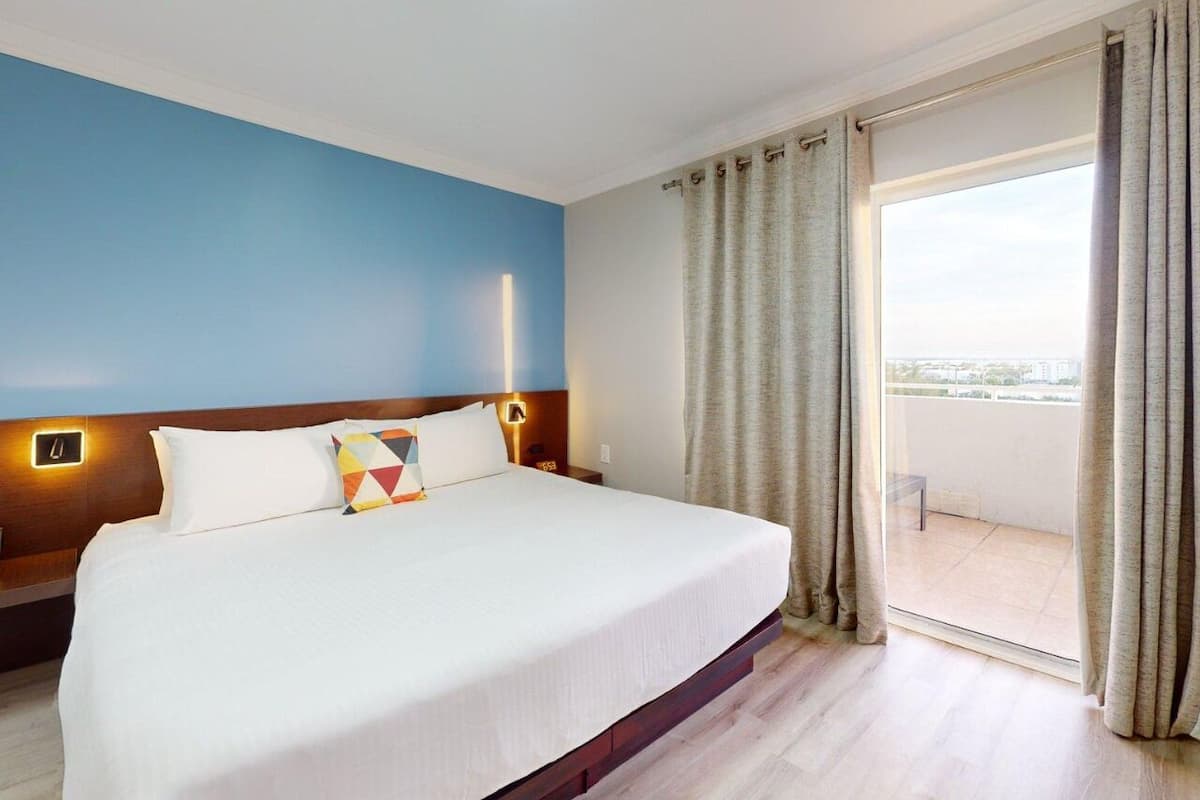 A hotel room with a large bed, a nightstand, a wall-mounted lamp, and a view through a glass door.