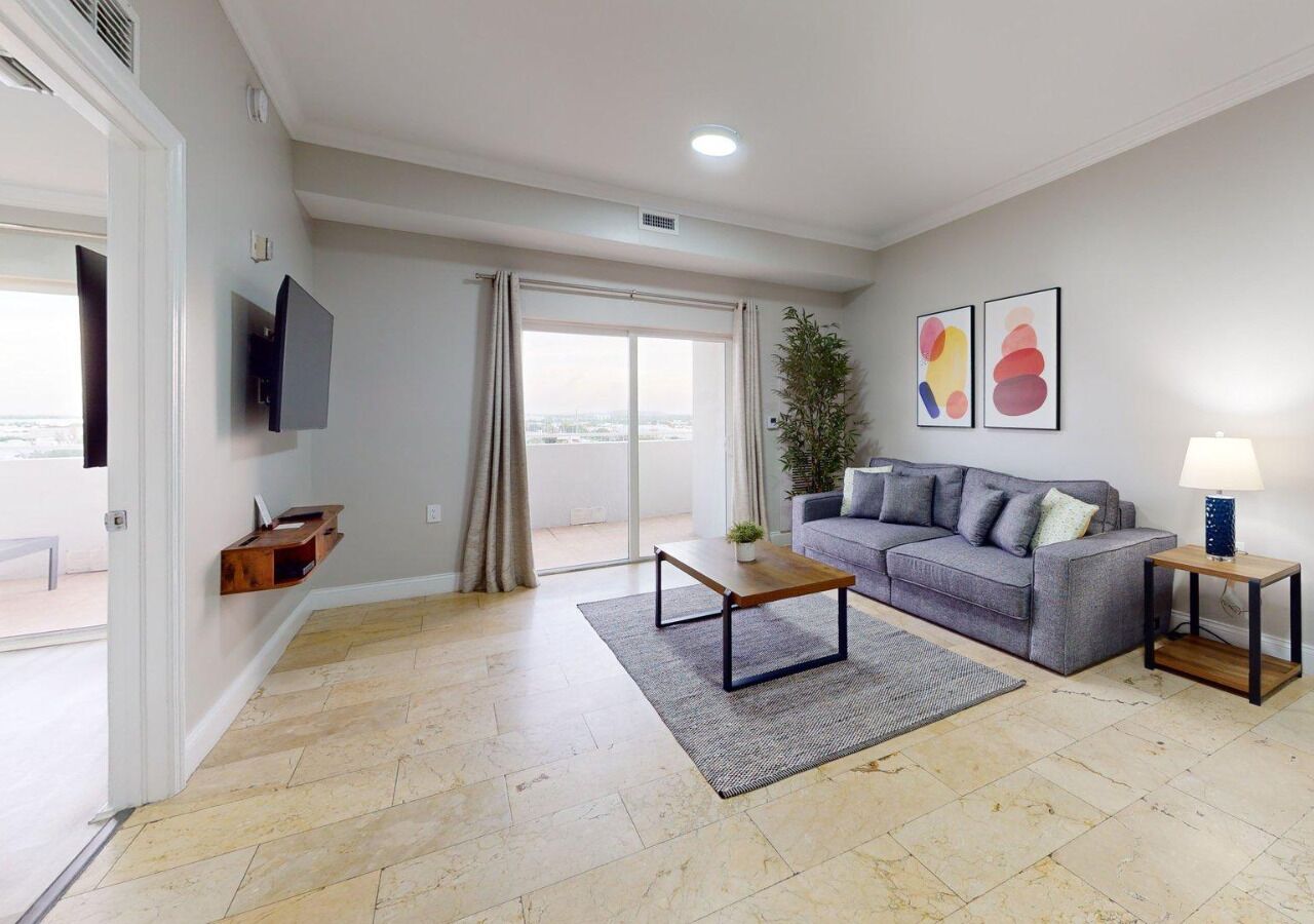 A modern living room with a grey sofa, a wooden coffee table, a small side table with a lamp, a flat-screen TV mounted on the wall, and a large window with curtains.