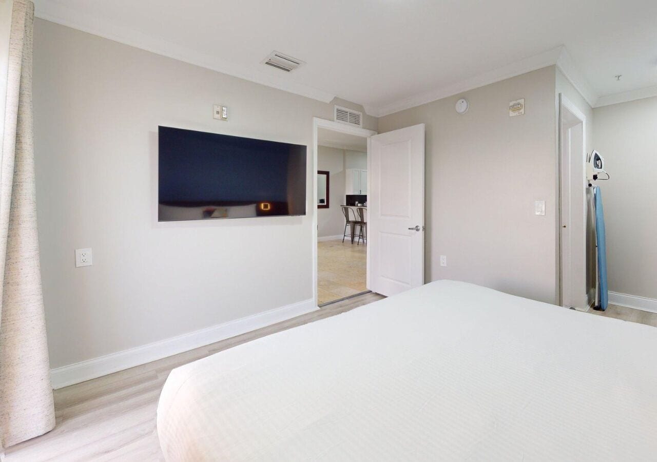 Habitación de hotel con una cama grande, un televisor de pantalla plana montado en la pared, una puerta que da a un baño y una zona de kitchenette visible.