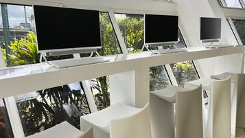 A modern office with three computer monitors on a white desk, large windows with a view of greenery, and white chairs.