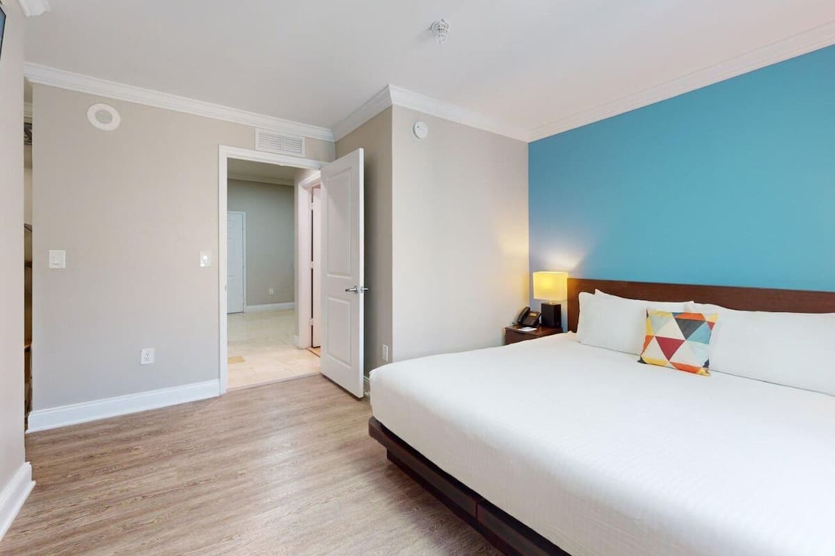 A hotel room with a bed, a nightstand, a lamp, and a door leading to another room.