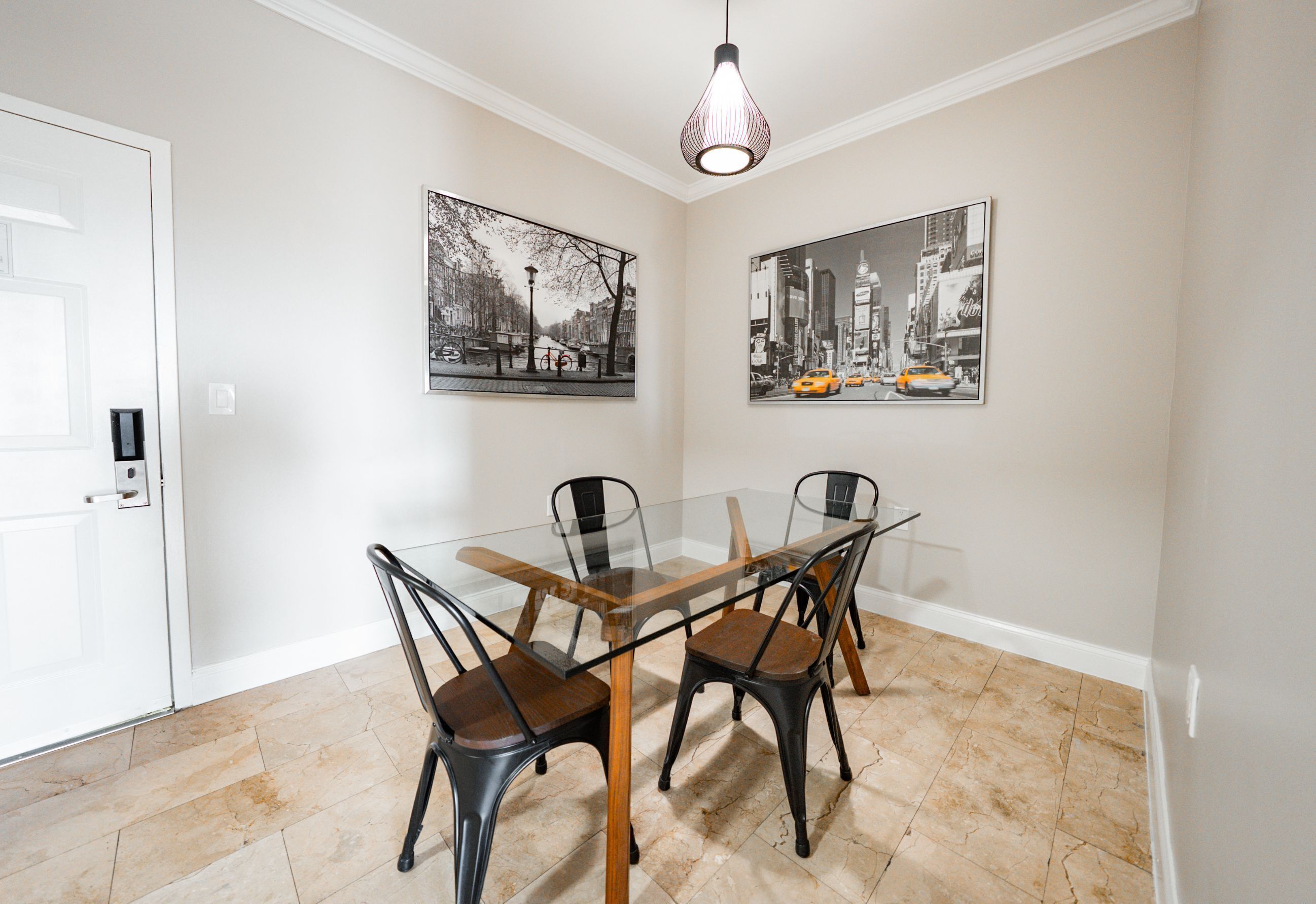 Uma sala de jantar com mesa de vidro e quatro cadeiras, duas pretas e duas castanhas, com uma porta branca e duas imagens da paisagem urbana em preto e branco nas paredes.