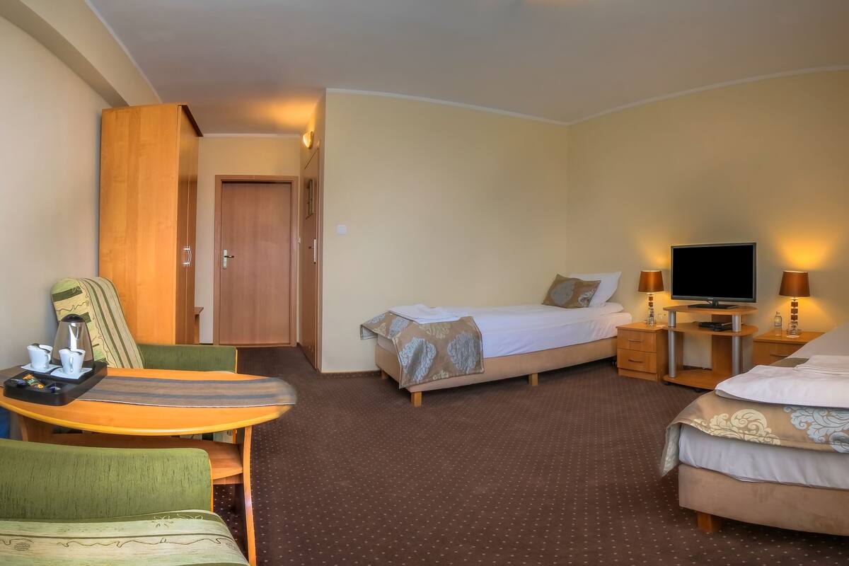 A hotel room with two beds, a TV, a desk with a chair, and a small table with a kettle and cups.