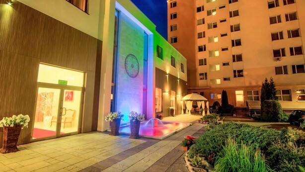 A hotel entrance with a modern facade, a visible clock, and a well-lit outdoor area with a fountain and potted plants. - Confero (Nysa)