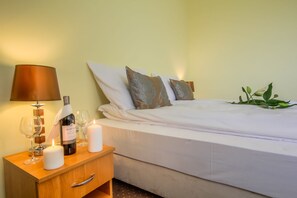 A neatly made bed with white linens, two decorative pillows, a bedside lamp, a bottle of wine, and two lit candles on a wooden nightstand.
