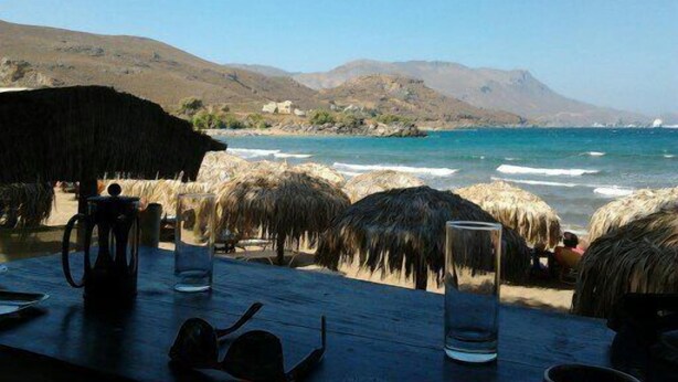 Un restaurant en bord de mer avec des tables en bois, des parasols en chaume et une vue sur l’océan et les collines.