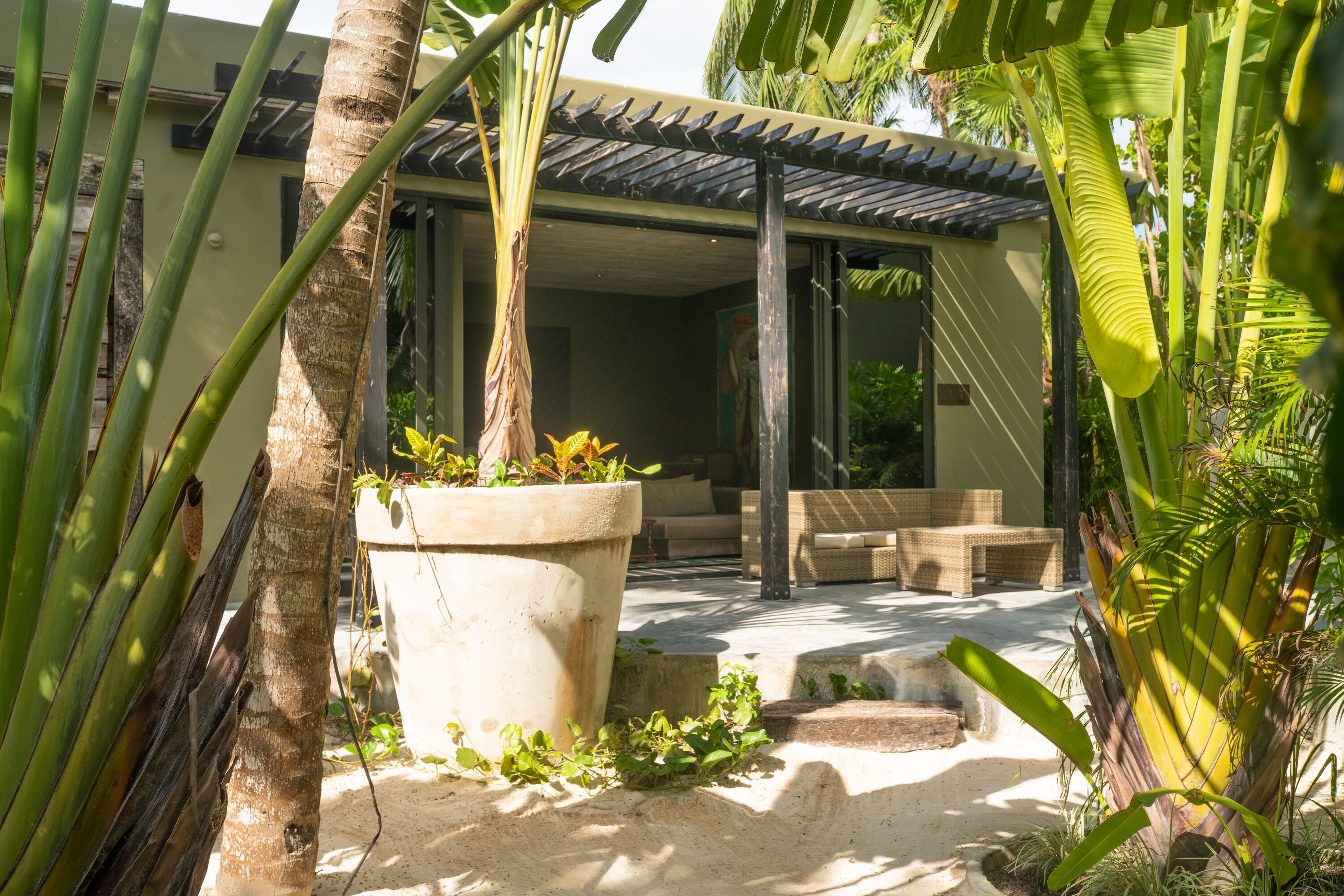 A covered outdoor seating area with a concrete floor, surrounded by palm trees and large potted plants.