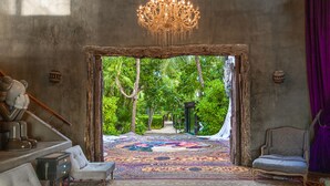 A room with a large, ornate chandelier, a patterned carpet, and a view of a lush garden through an open doorway. - Casa Malca (Tulum)