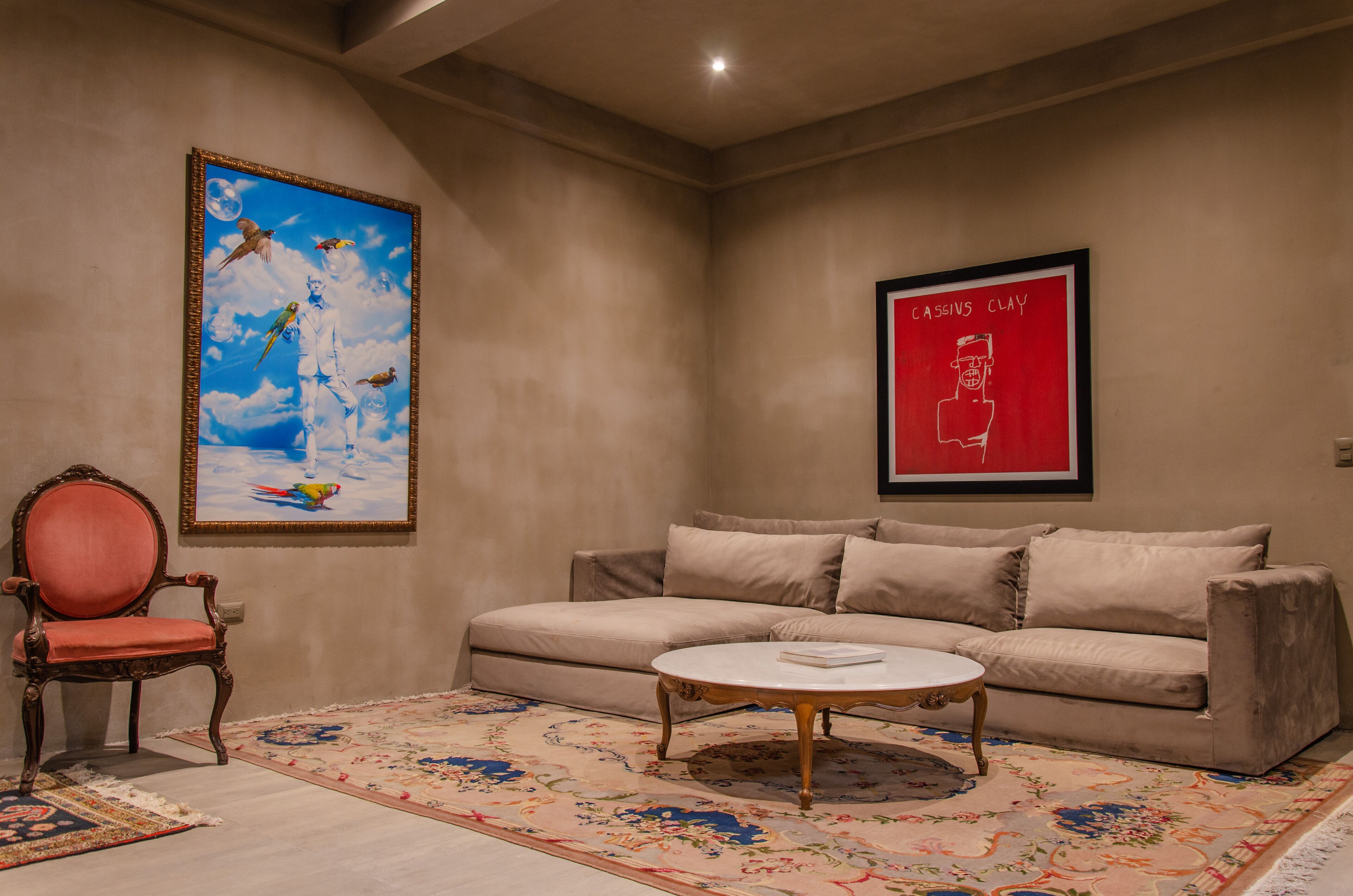 Una habitación con un sofá color crema, un sillón rojo, una mesa de centro redonda y dos obras de arte enmarcadas en las paredes.