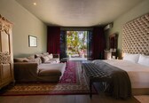 A spacious bedroom with a large bed, a sofa, and a bench. There is a view of palm trees through the window.